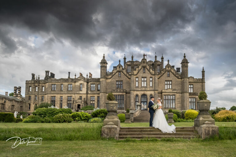 Bride and groom standing on the steps outside of Allerton Castle, with dark grey storm clouds in the sky.
