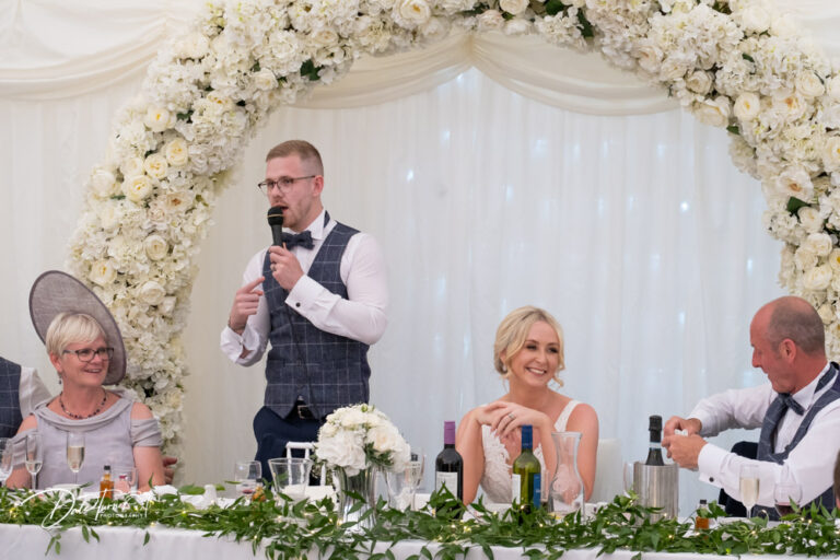 Groom giving a speech with his bride is sat next to him smiling. At The Oaks Golf Club.