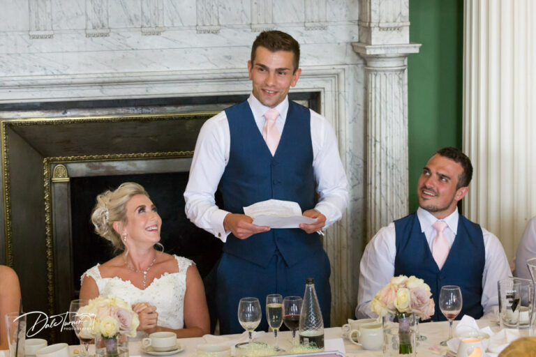 Groom giving a speech at Hazlewood Castle while the bride and the best man look at him and smile.
