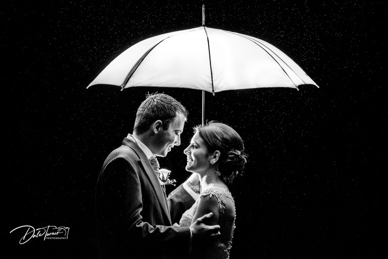Bride and groom with a white umbrella in the rain at night time.