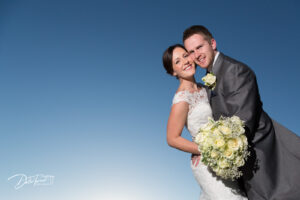 Bride and groom standing cheek to cheek and smiling with a clear blue sky behind them.