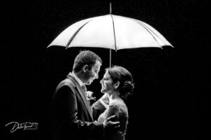 Twilight photograph of a bride and groom looking at each other under an umbrella in the rain.