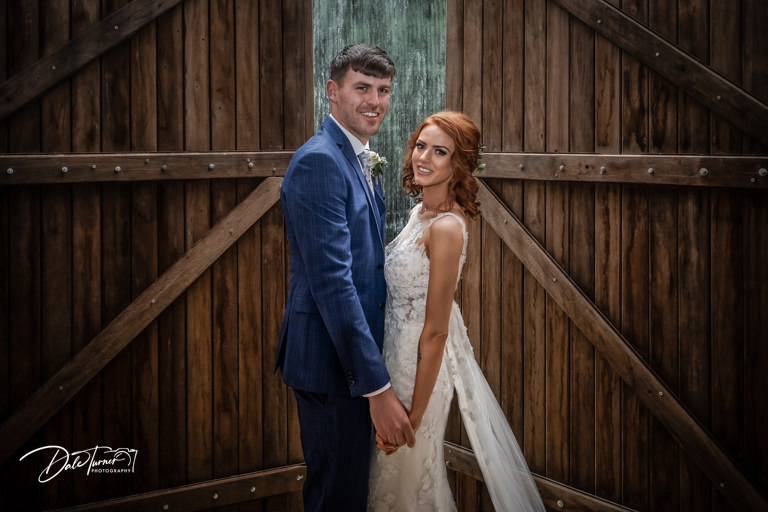 Bride and groom standing in front of large wooden door and smiling, at The Yorkshire Barns.