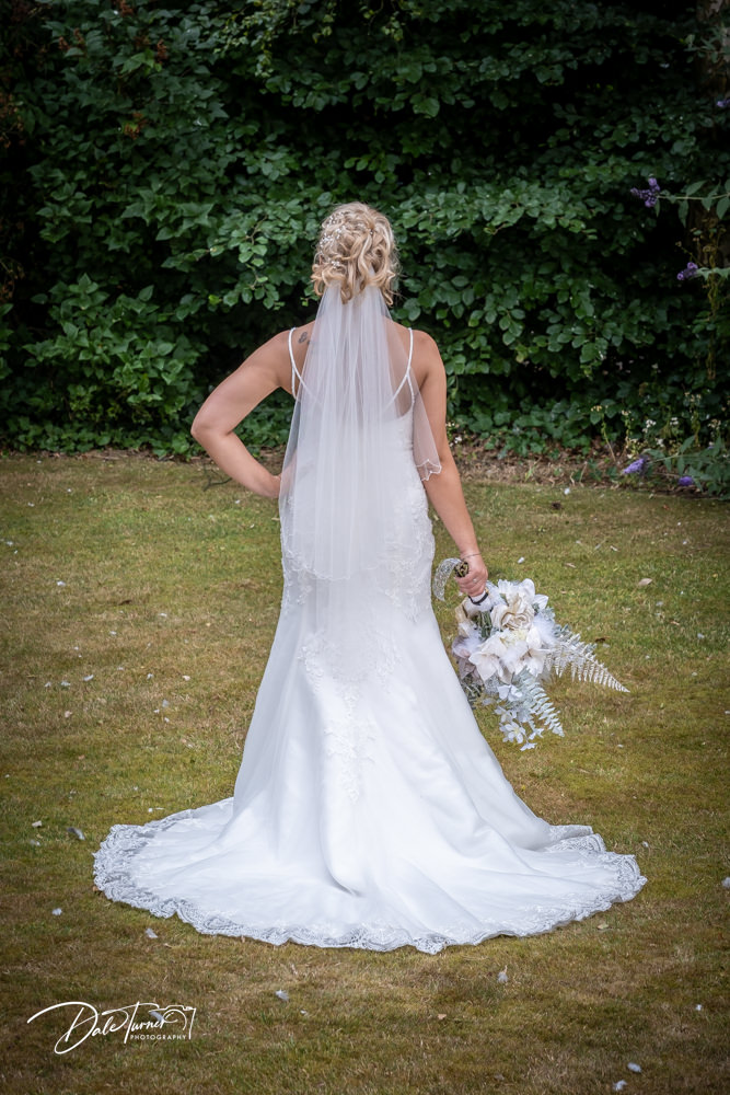 The back of a bride, standing with her bouquet and a hand on her hip. In the grounds of The Parsonage, Escrick.
