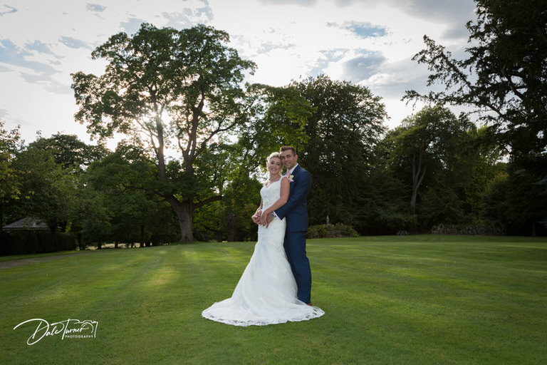 Groom hugging bride from behind as the sun shines through the trees in the grounds of Hazlewood Castle.