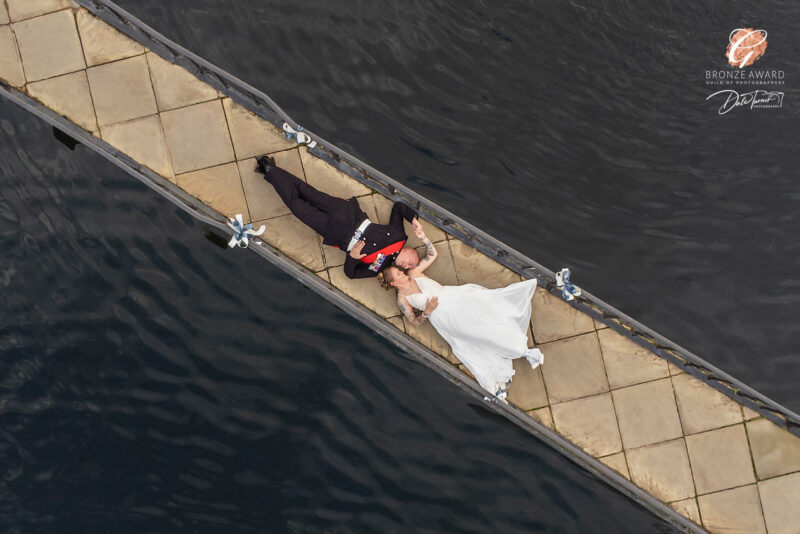 Award winning drone photograph of a bride and groom laying down on a bridge and holding hands, at Waterton Park Hotel.