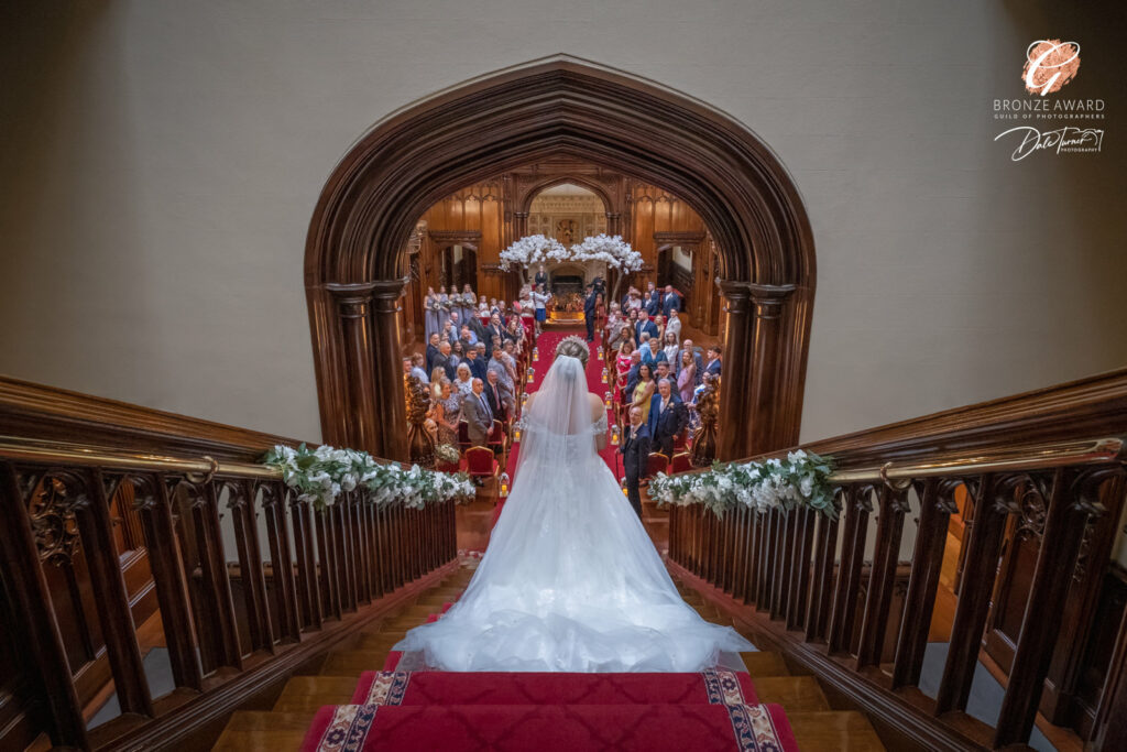 Award winning photograph of a bride (from behind) walking down the staircase inside Allerton Castle, making her way to the ceremony. Friends and family are watching while the groom waits at the end of the aisle.