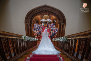 Award winning photograph of a bride (from behind) walking down the staircase inside Allerton Castle, making her way to the ceremony. Friends and family are watching while the groom waits at the end of the aisle.
