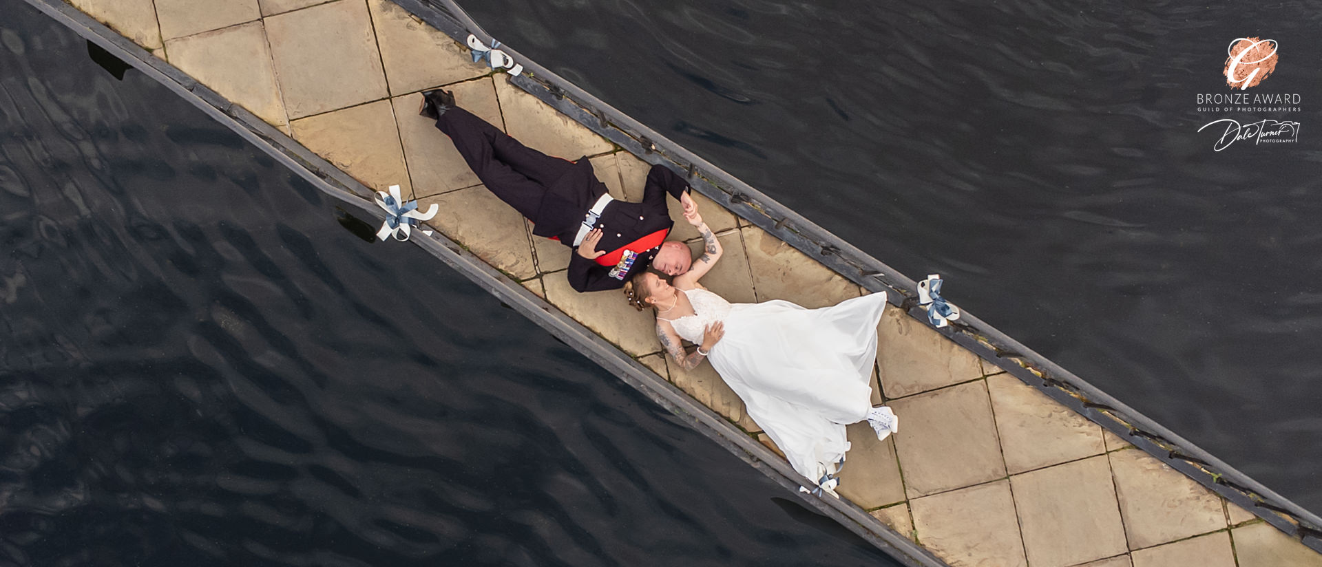 Bride and groom laying down on a bridge, holding hands. Taken from above with a drone at Waterton Park Hotel.