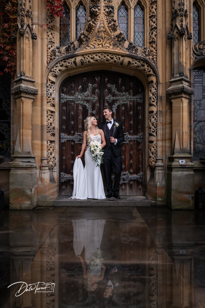 A serene bride and groom stand by the ornate main entrance of Carlton Towers, with the bride in a sleek white gown holding a bouquet and the groom in a classic black tuxedo. The intricate gothic architecture and the wooden doors create a romantic, timeless atmosphere, which is artfully reflected on the glossy surface below them, offering a hint of the venue's elegance.