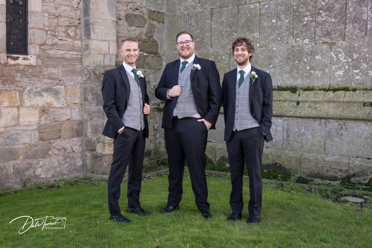 Three groomsmen smiling outside historical building.