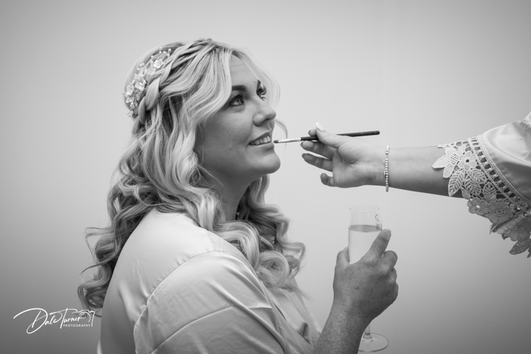 Bride having makeup applied before wedding, holding champagne.