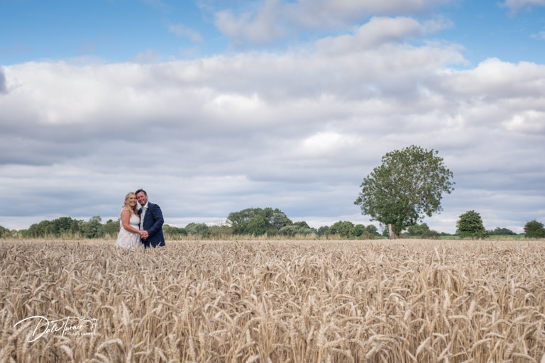 Bride and groom embracing in wheat field under cloudy sky.
