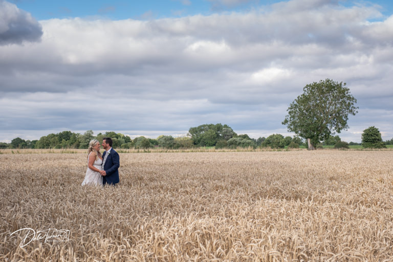 Bride and groom looking at each other embracing in wheat field under cloudy sky.