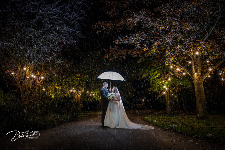 Bride and groom under an umbrella in the rain at night with fairy lights in the trees, at The Parsonage.