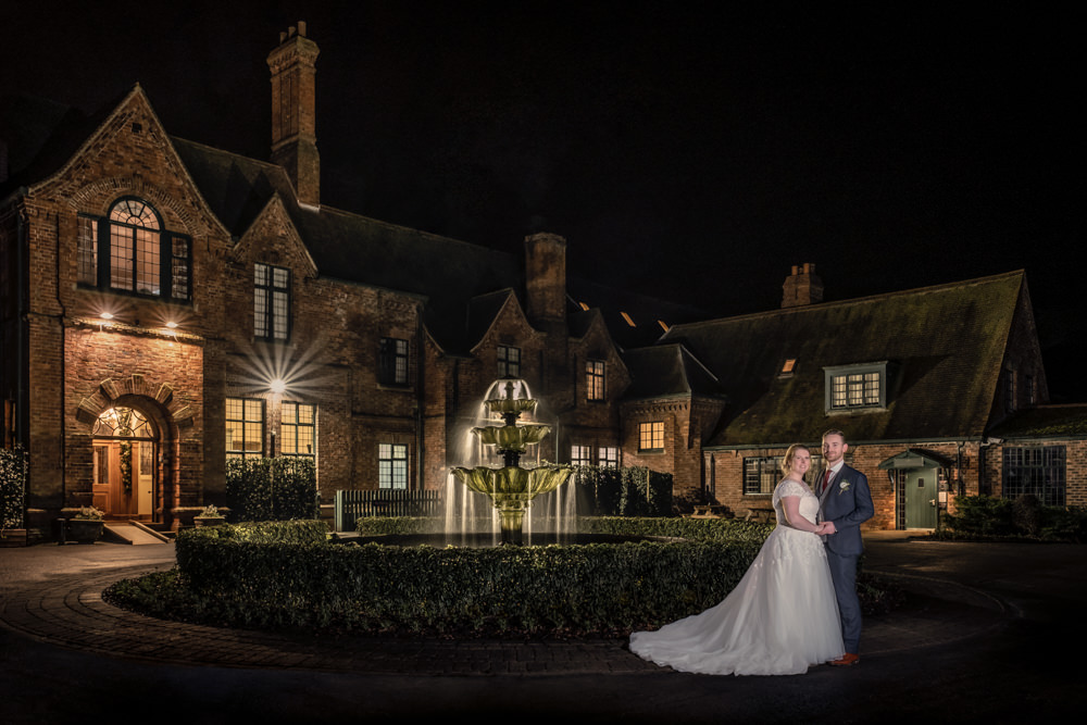 Newlyweds posing at night by illuminated manor and fountain.