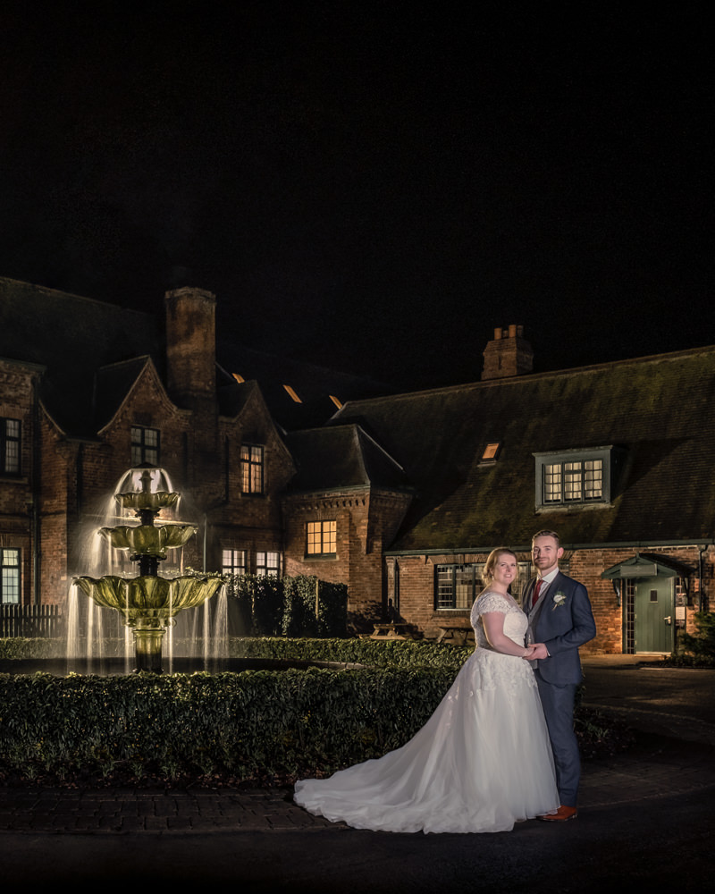 Newlyweds standing by fountain at night, historic house backdrop