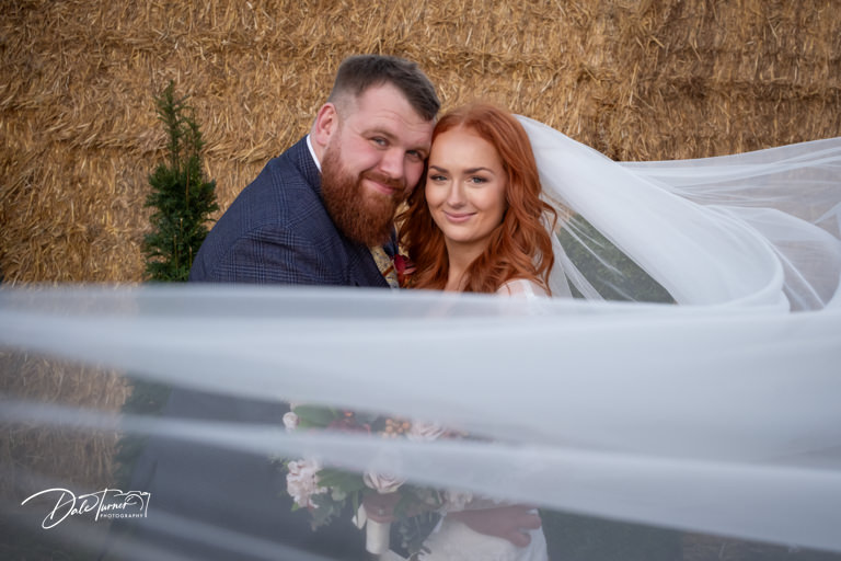 Bride and groom smiling behind sheer veil near hay
