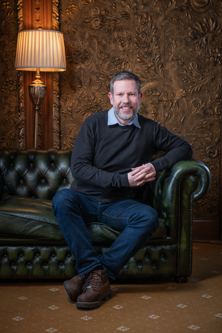Dale Turner smiling on green leather sofa, ornate background